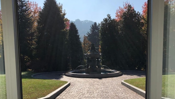 Un sendero pavimentado que lleva a una fuente, rodeada de árboles y un cielo despejado.
