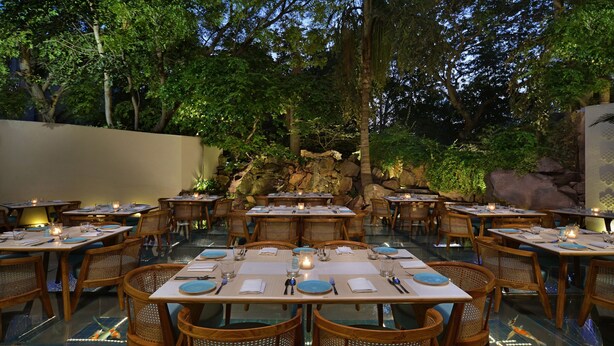 An outdoor dining area with tables set for multiple guests, surrounded by lush greenery and a rocky backdrop.