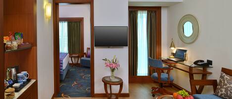 A hotel room with a bed, a TV, a desk with a computer, a chair, a small table with a vase of flowers, and a shelf with snacks and a coffee maker.