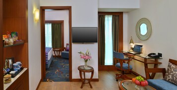 Habitación de hotel con cama, televisión, escritorio con ordenador, silla, mesita con un florero y estante con aperitivos y cafetera.