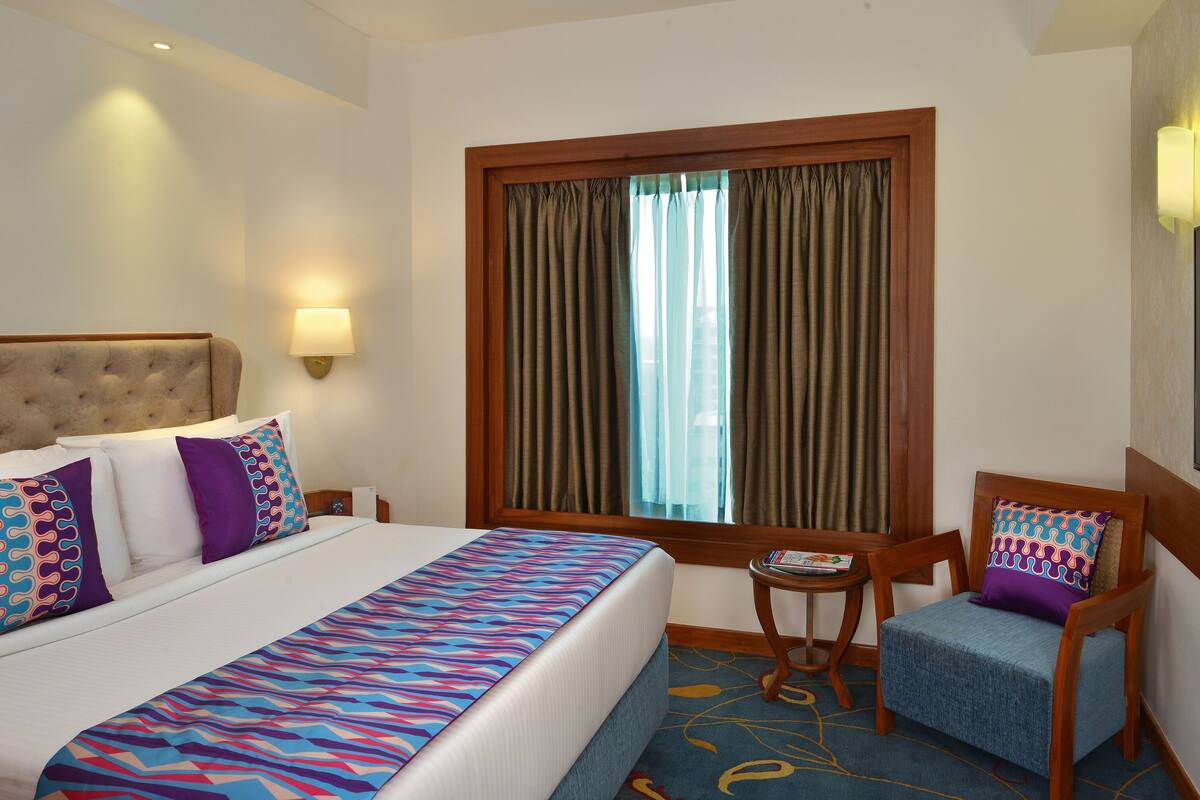 Habitación de hotel con cama, una silla, una mesita, televisor y ventana con cortinas.
