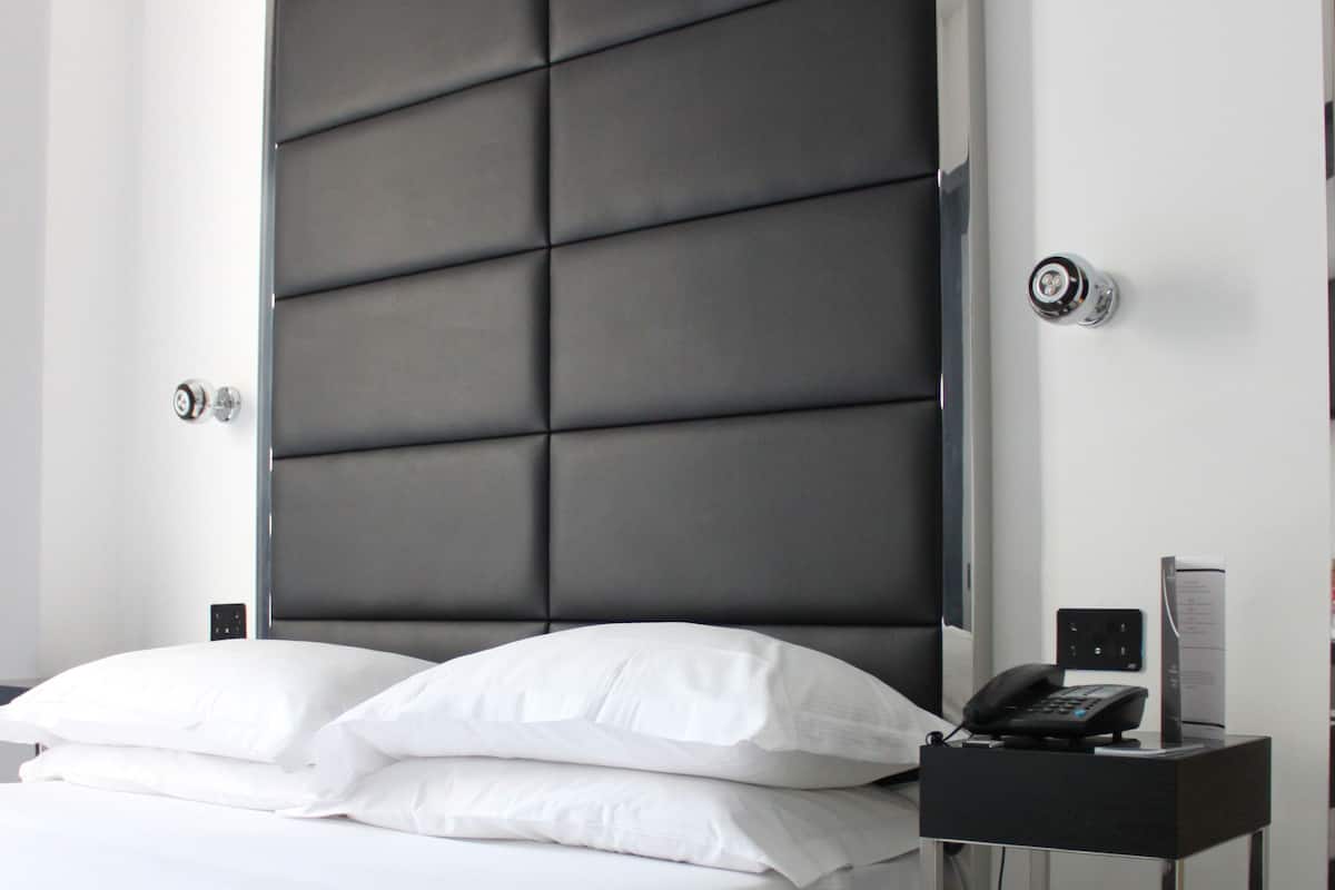 A modern hotel room with a large bed, a bedside table with a phone and a small lamp, and a wall-mounted thermostat.