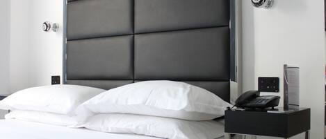A modern hotel room with a large bed, a bedside table with a phone and a small lamp, and a wall-mounted thermostat.