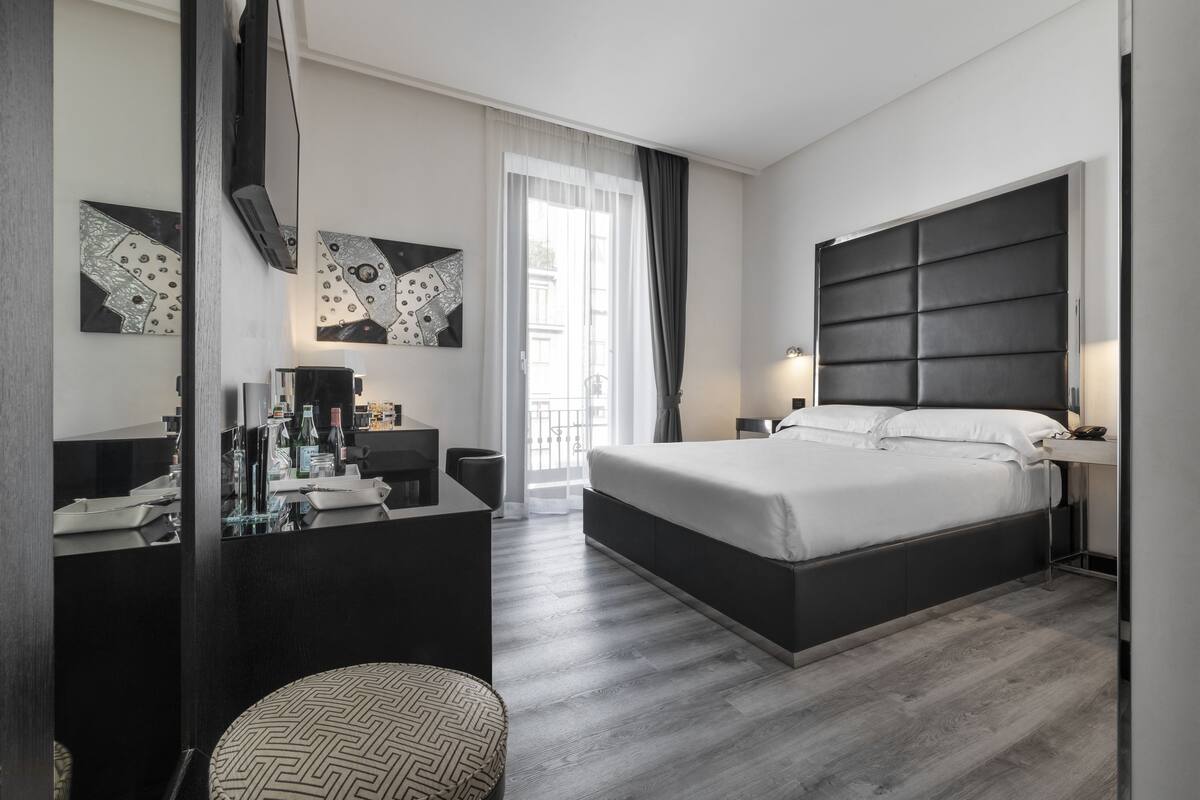 A modern hotel room with a large bed, a desk with a chair, and a vanity with a mirror.