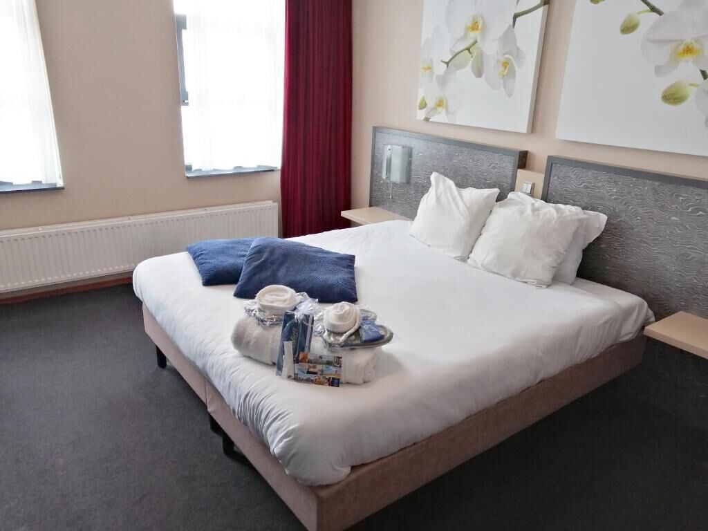 Un cuarto de hotel con una cama grande, una mesita de noche, una cortina roja y dos cuadros con orquídeas en la pared.