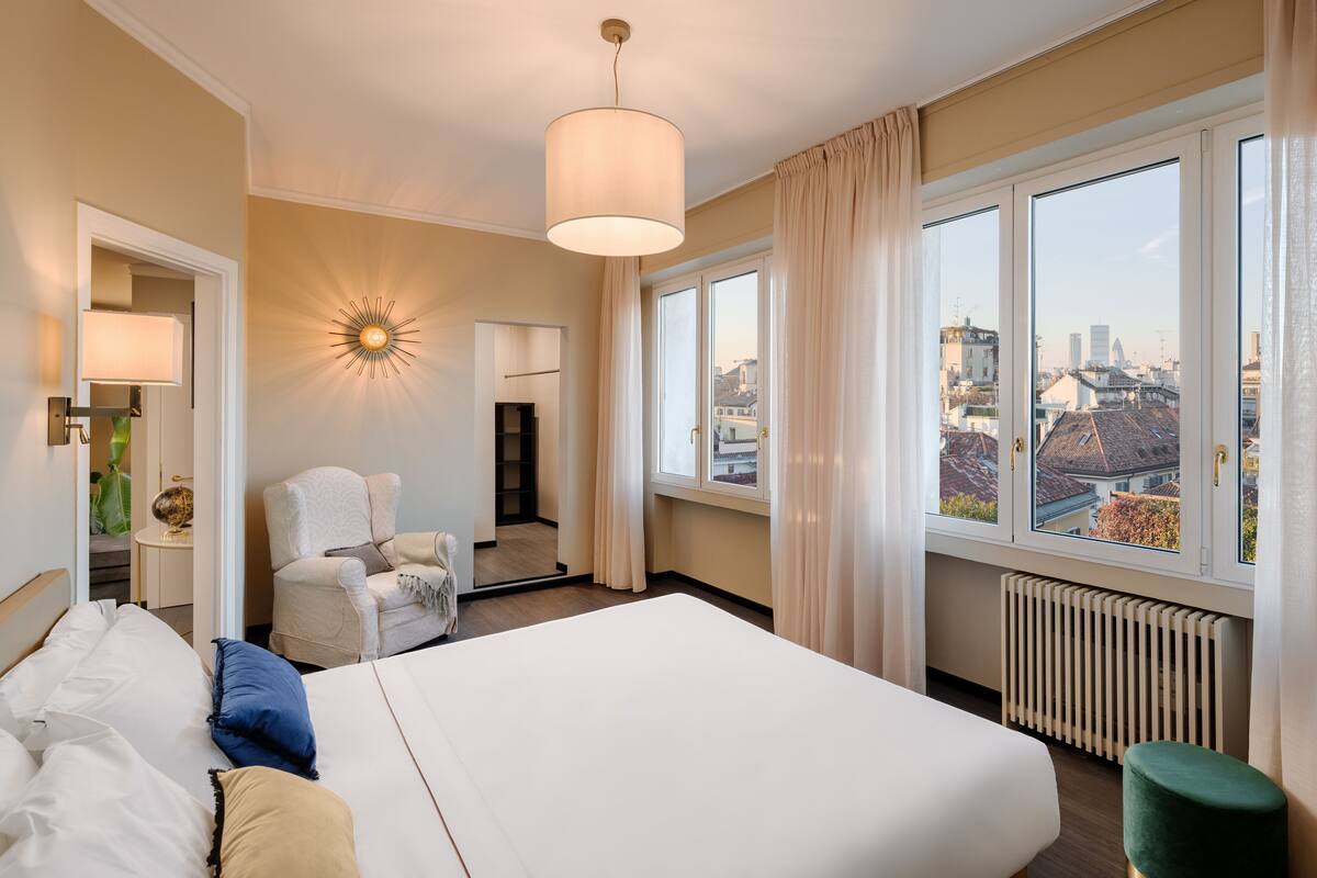 A hotel room with a large bed, a chair, a desk, and a view of the city through the windows.