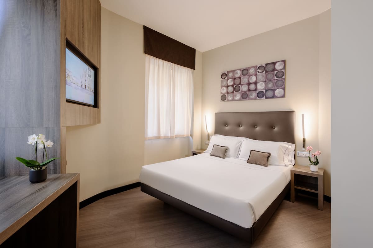 A modern hotel room with a large bed, a bedside table with a lamp, a wall-mounted TV, and a decorative wall piece.