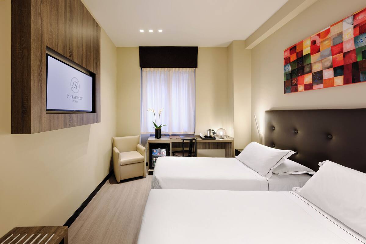 A hotel room with two beds, a desk, a chair, a TV, and a colorful artwork on the wall.