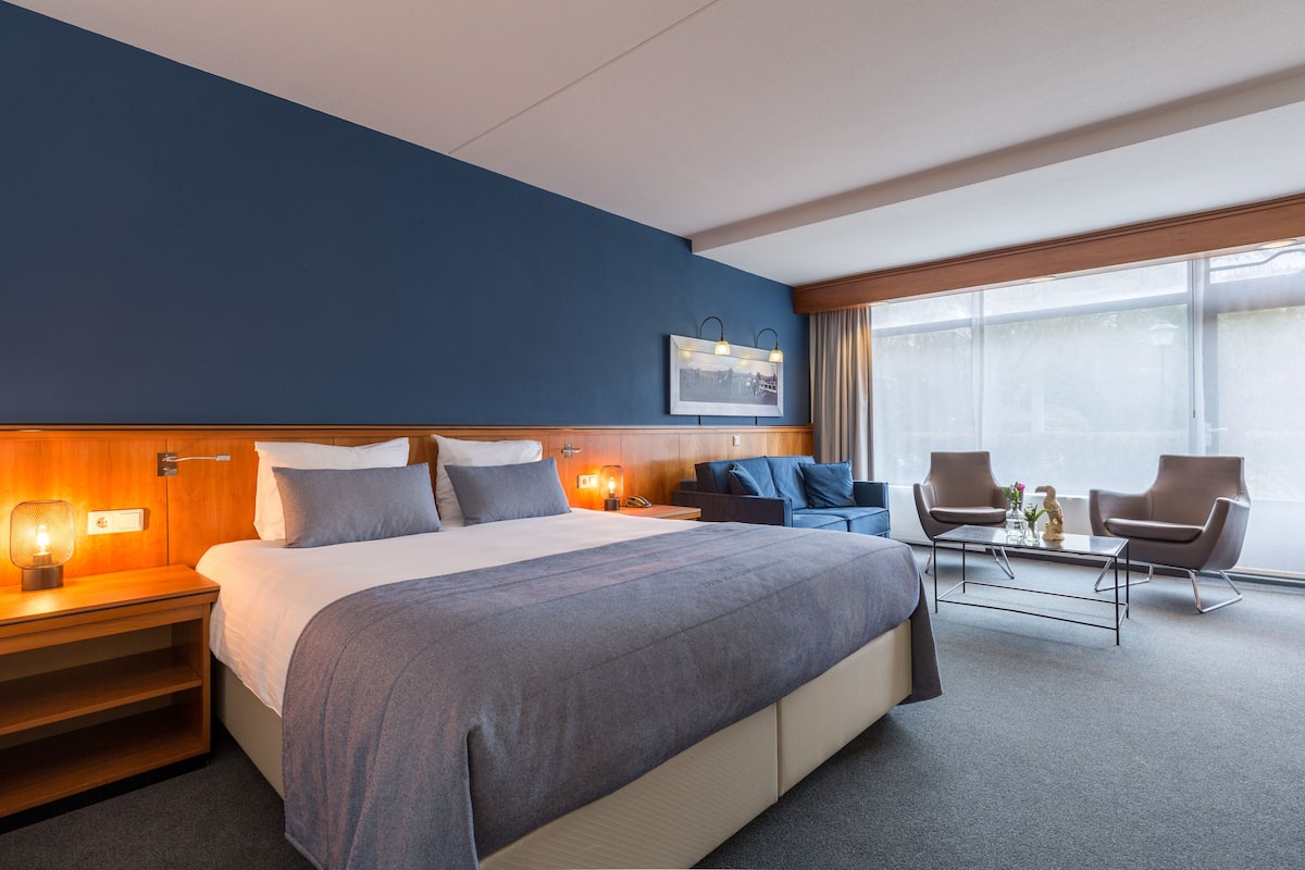 Ein Hotelzimmer mit einem großen Bett, Nachttischen, einem Schreibtisch, zwei Sesseln und einem Fenster mit Vorhängen.