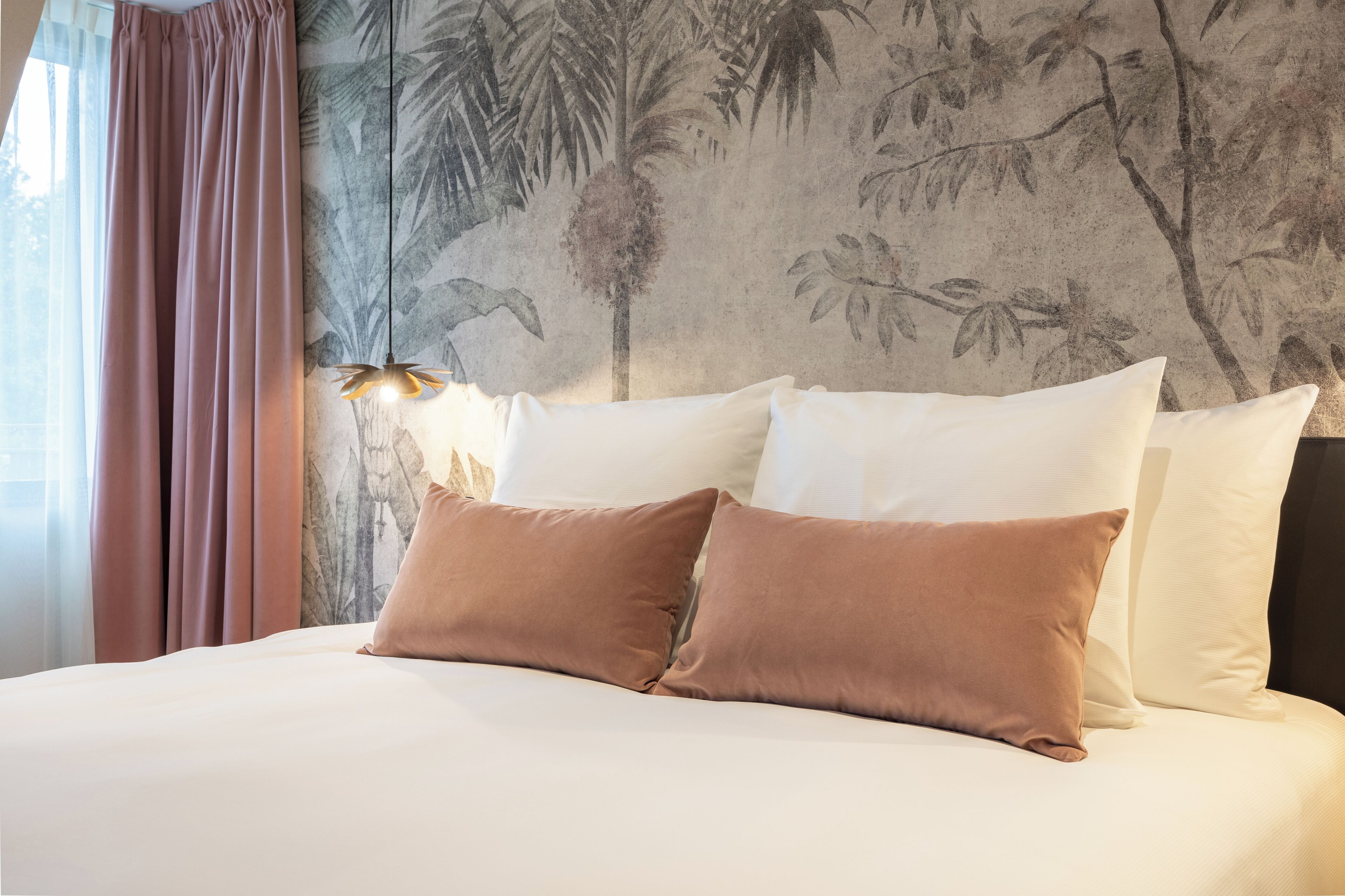 Een netjes opgemaakt bed met witte en bruine kussens, een muur met een tropisch muurschildering, en een raam met roze gordijnen.