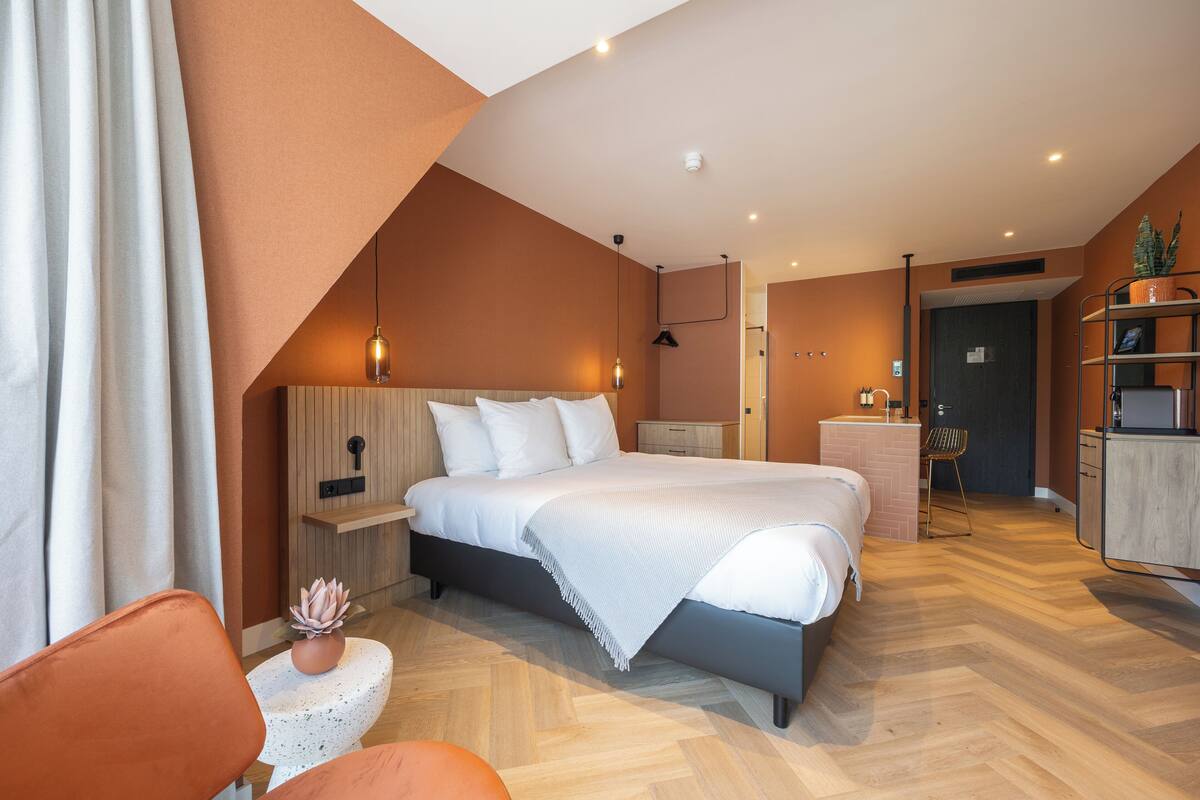 Ein modernes Hotelzimmer mit einem großen Bett, einem kleinen Schreibtisch, einem Sessel und einer Pflanze.