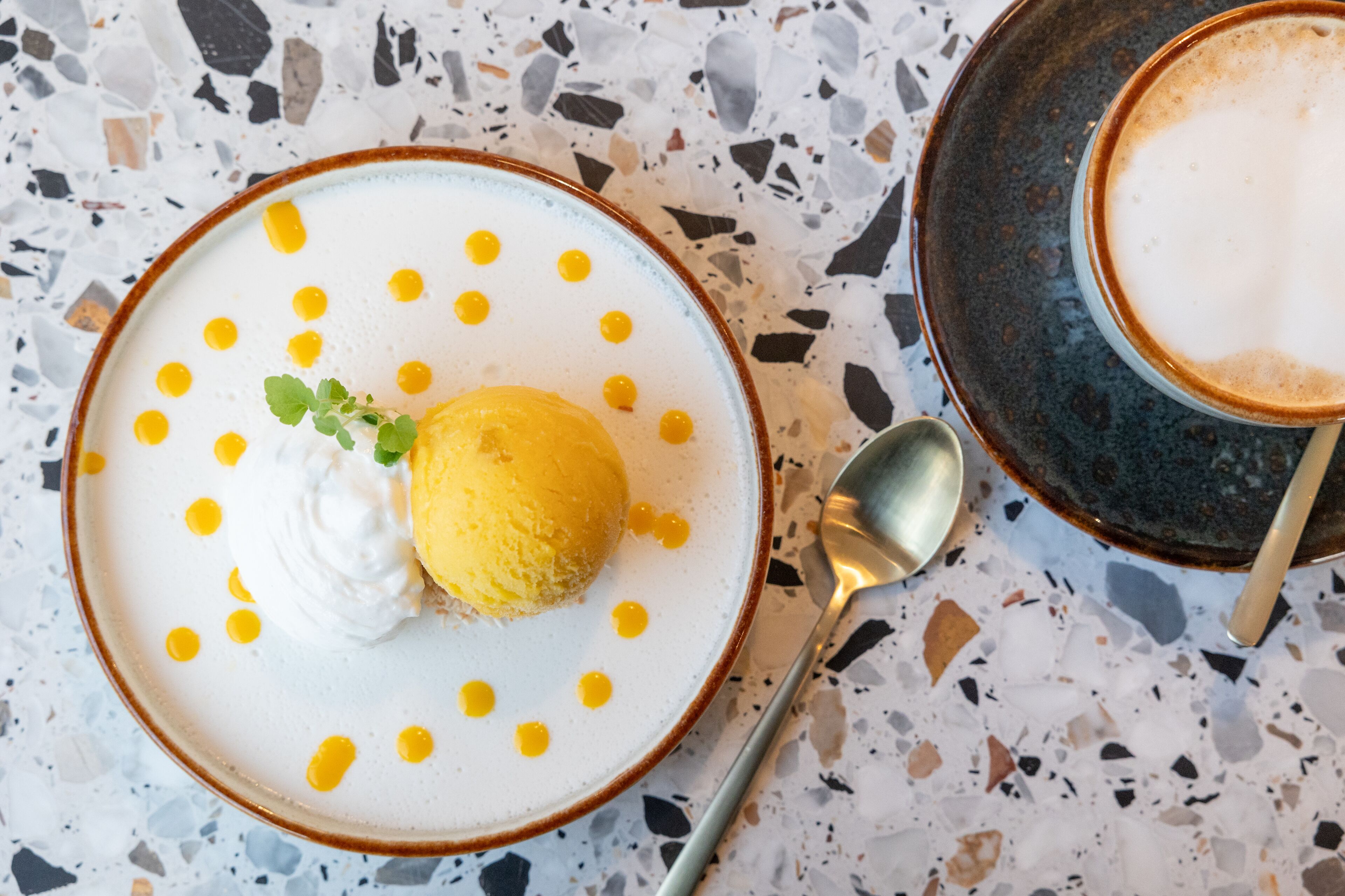 A dessert plate with a scoop of ice cream and whipped cream, garnished with yellow dots and a mint leaf, on a speckled countertop.