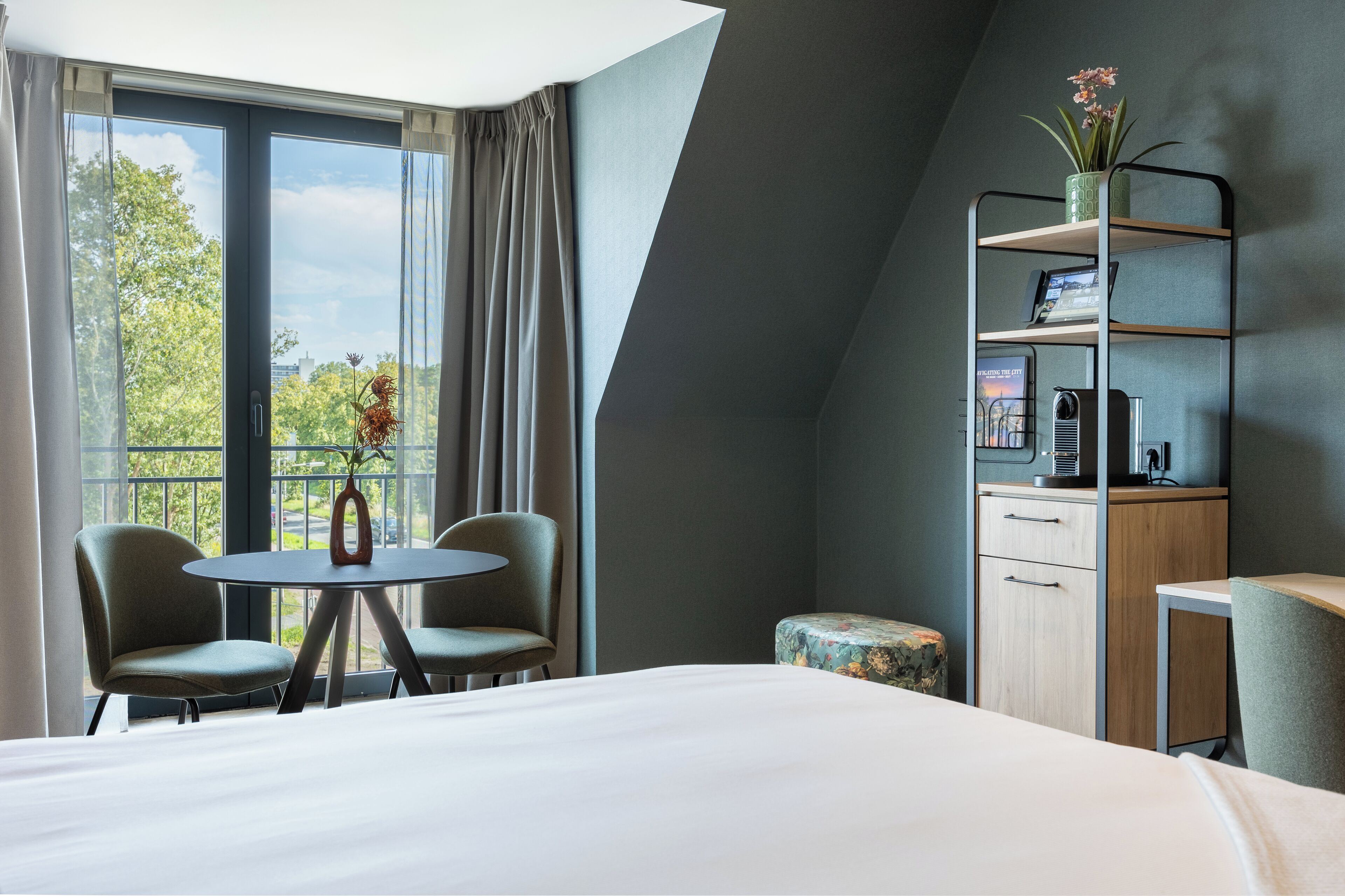 Ein Hotelzimmer mit einem Bett, zwei Sessel, einem kleinen Tisch mit einer Vase, einem Schreibtisch mit einem Computer und einem Bücherregal.