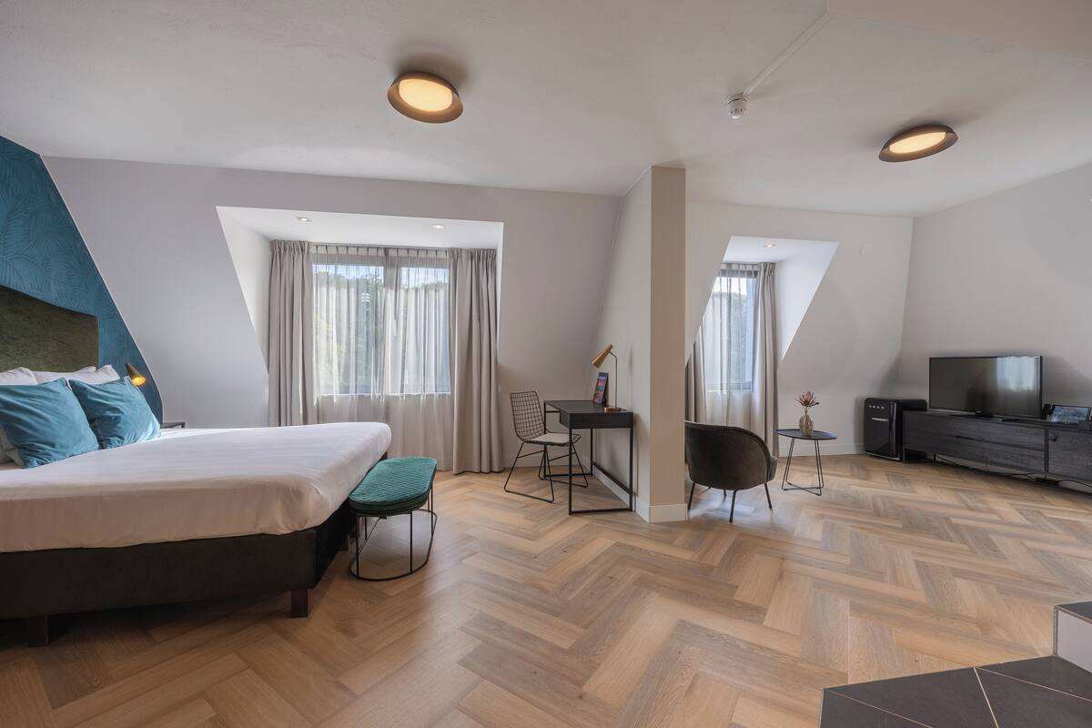 Ein modernes Hotelzimmer mit Bett, Schreibtisch, Sessel, Fernseher und einem kleinen Tisch mit einer Vase.