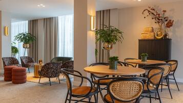 Un moderno vestíbulo de hotel con mesas redondas y sillas de mimbre, una zona de estar con sofás y plantas decorativas.