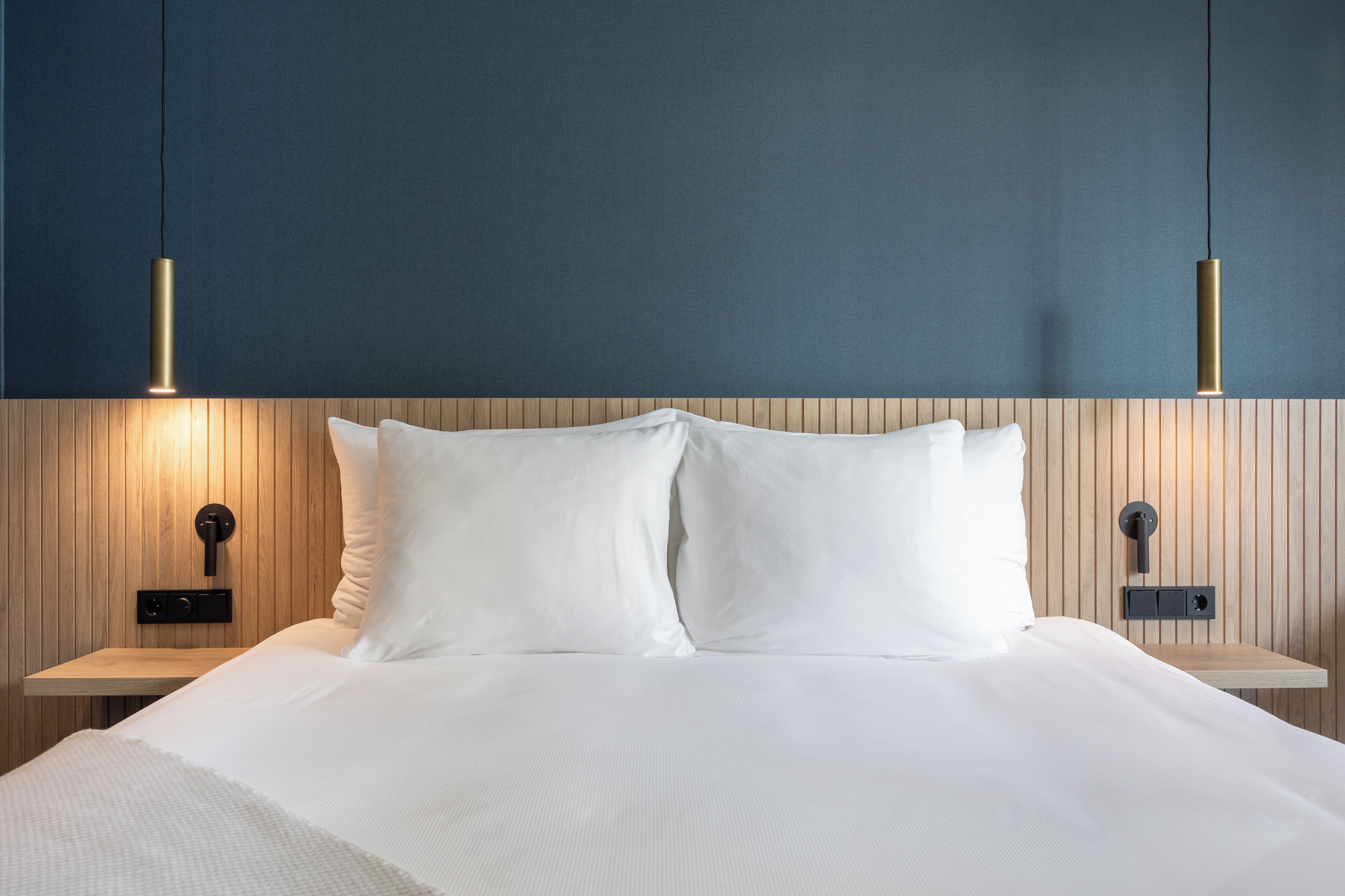 Een netjes opgemaakt bed met witte lakens, twee nachtkastjes met wandlampen en een houten hoofdbord.