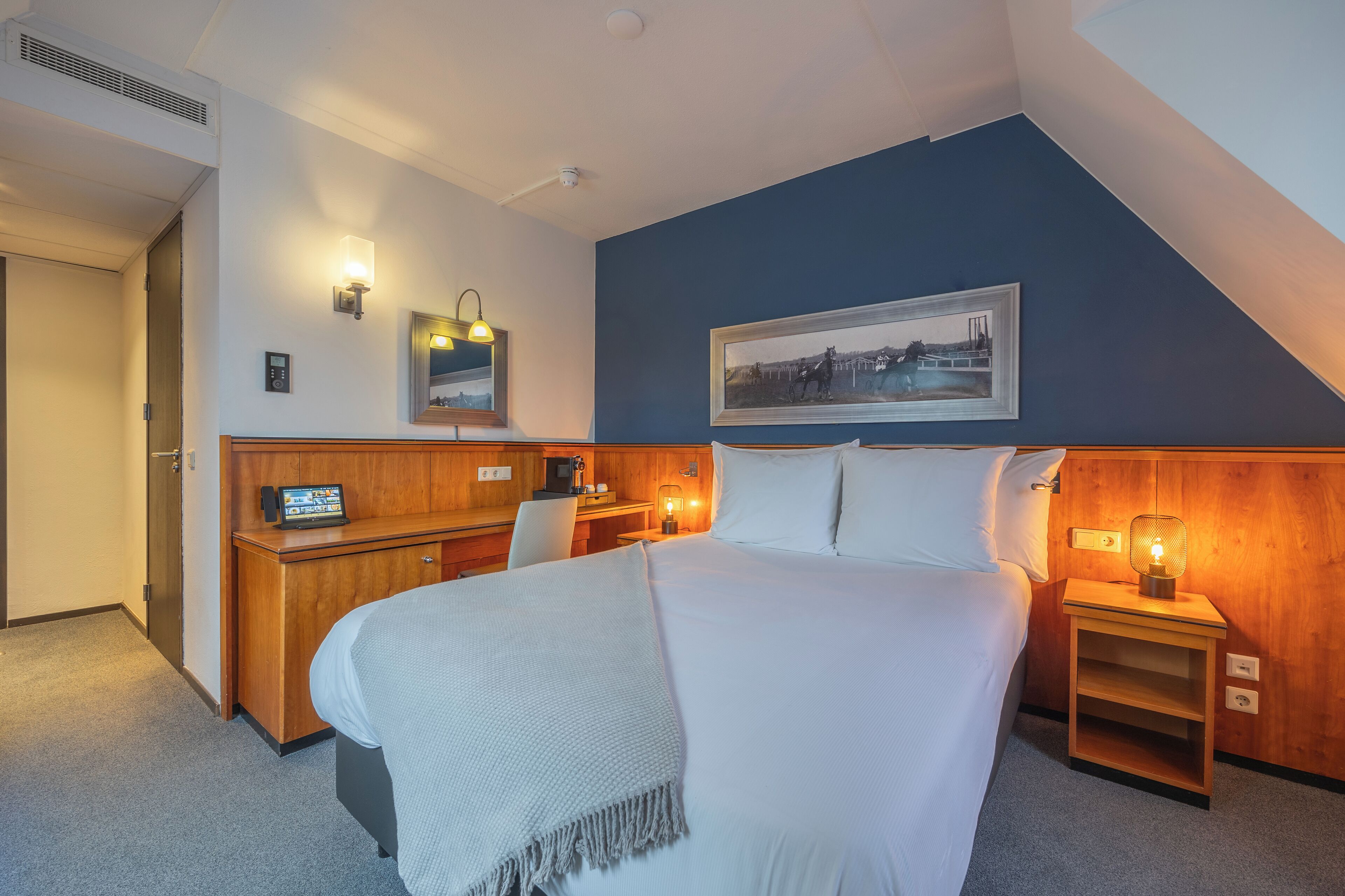 Quarto de hotel com cama, mesas de cabeceira, escrivaninha e uma cadeira. Há luminárias na parede e um quadro pendurado.