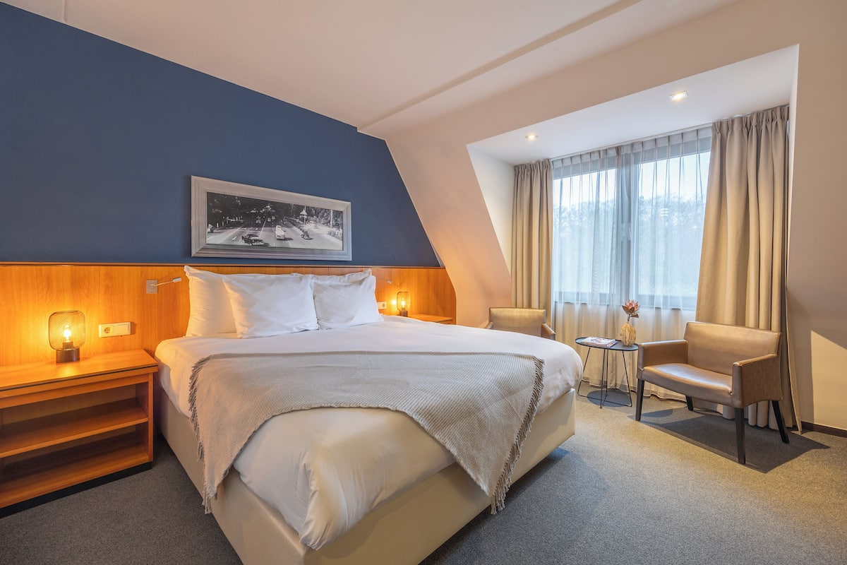Ein Hotelzimmer mit einem großen Bett, Nachttischen mit Lampen, einem Sessel, einem kleinen Tisch mit einer Vase und einem Fenster mit Vorhängen.