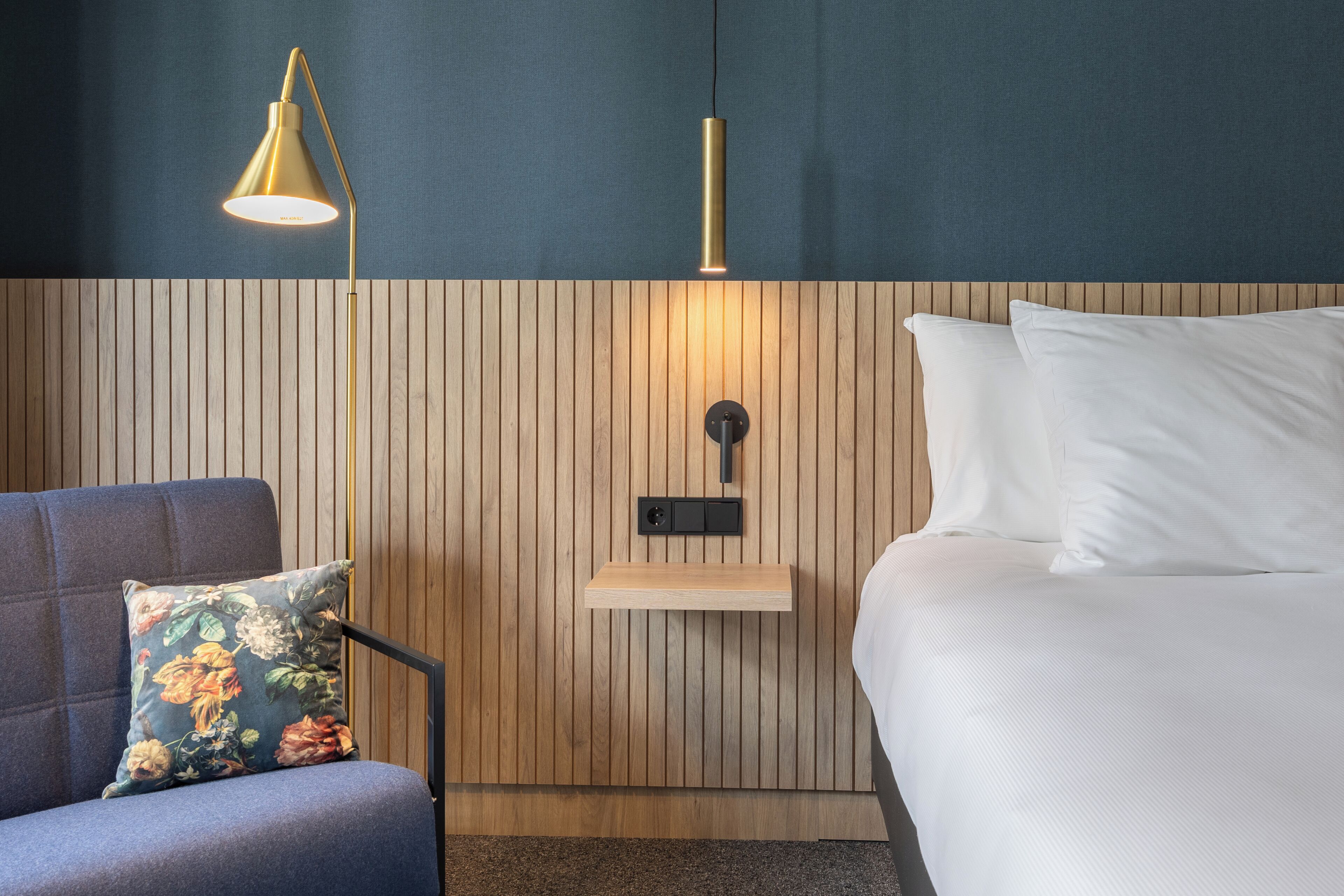 Une chambre d’hôtel avec un mur lambrissé en bois, un lit avec du linge de lit blanc, un fauteuil bleu avec un coussin à motifs floraux et une lampe sur pied.