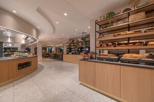 Uma área de café da manhã moderna com balcões de madeira, diversos tipos de pão e uma cozinha ao fundo.