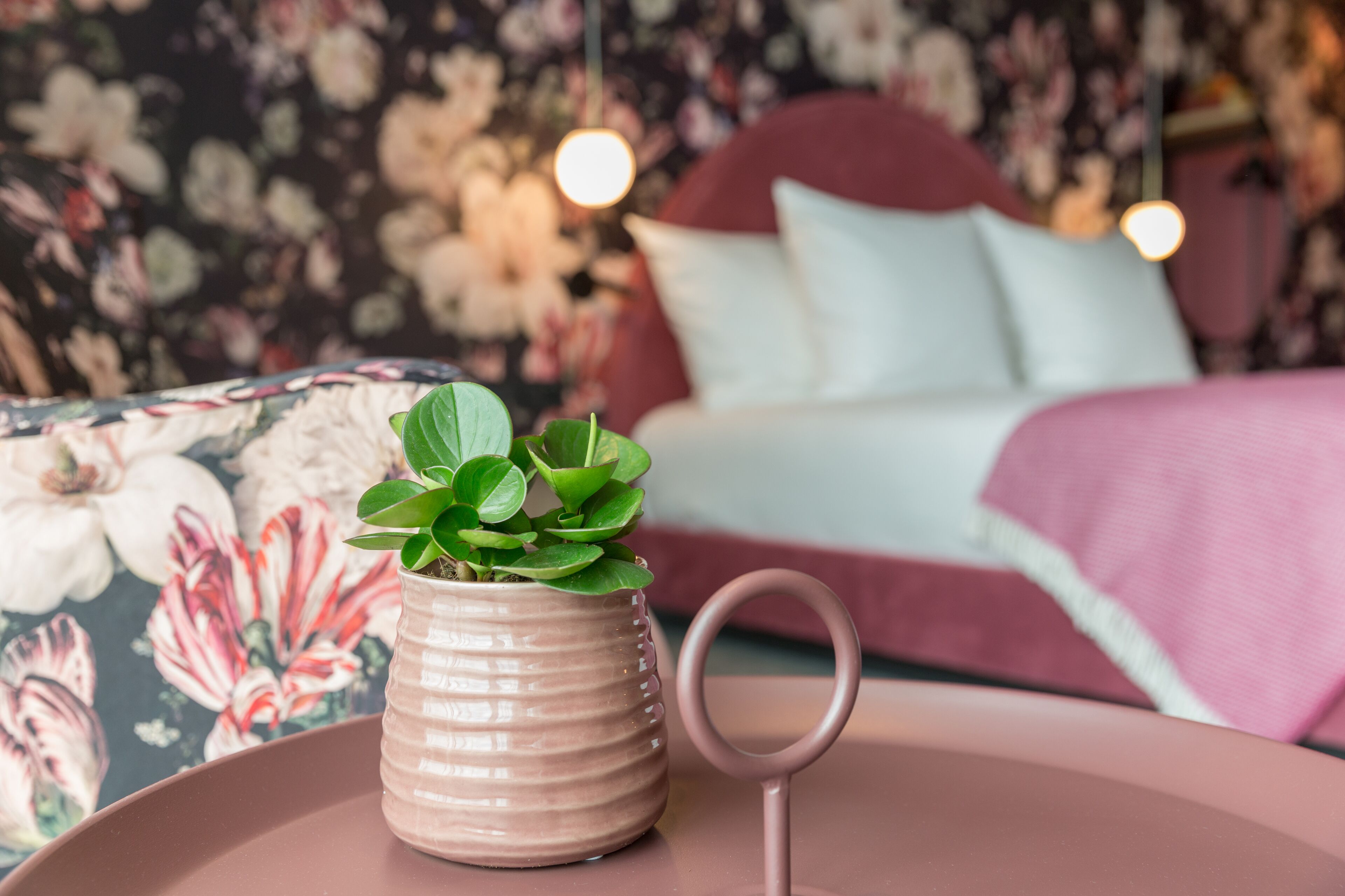 Een bank met bloemenprint, een potplant en een bed met een roze deken in een kamer met bloemenbehang en hangende lampen.