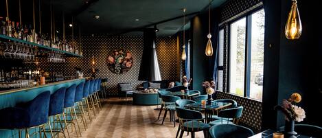 A bar area with a herringbone wooden floor, teal bar stools, and a variety of bottles and glasses on shelves.