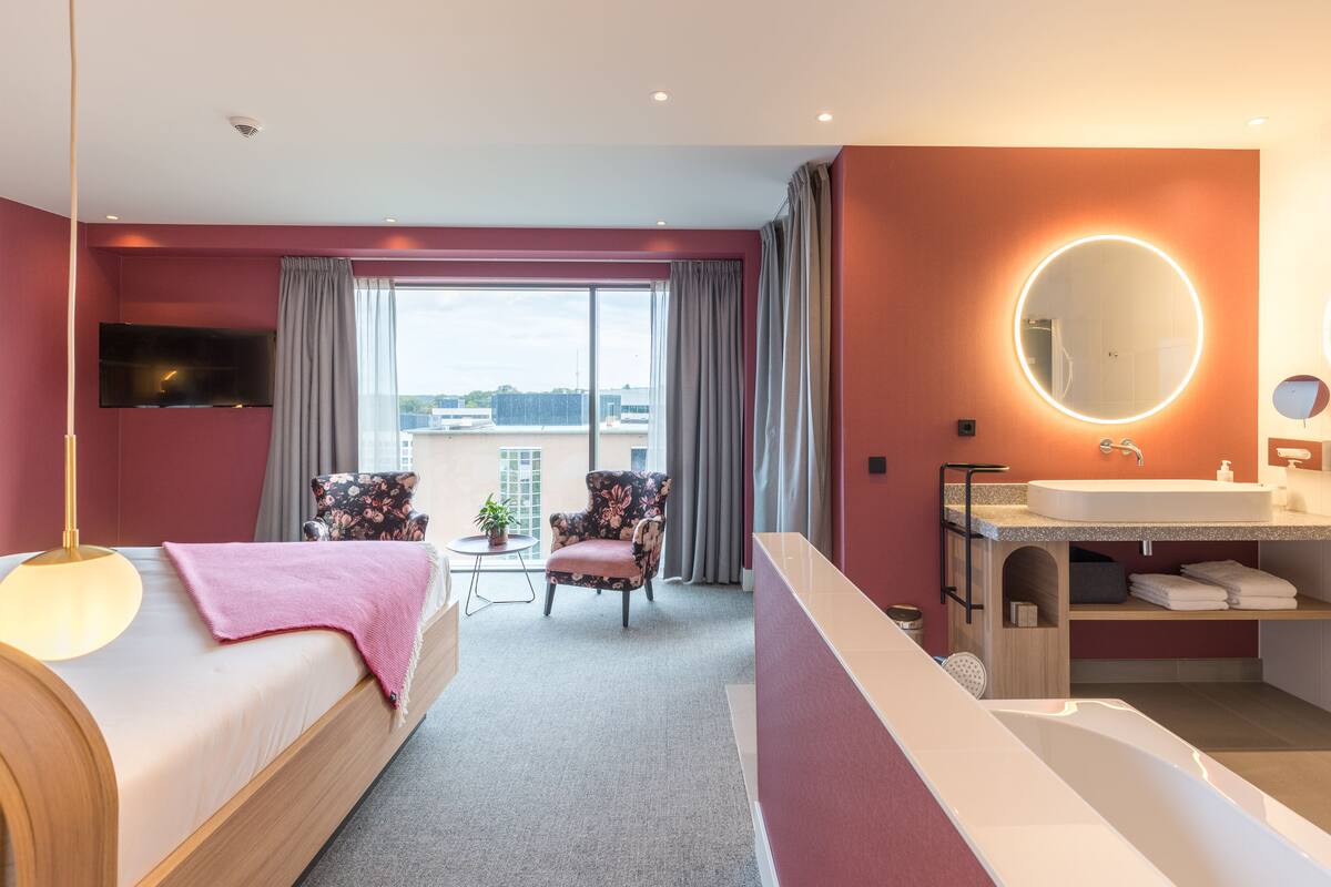 Une chambre d’hôtel moderne dotée d’un grand lit, d’un coin salon avec un fauteuil et une table, d’une salle de bain avec un lavabo et un miroir, et offrant une vue sur la ville grâce à de grandes fenêtres.