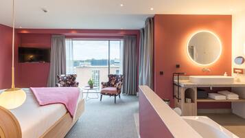 A modern hotel room with a large bed, a sitting area with a chair and table, a bathroom with a sink and mirror, and a view of the city through large windows.