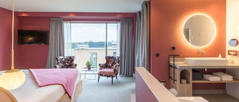 Una habitación de hotel moderna con una cama grande, un área de descanso con una silla y una mesa, un baño con lavamanos y espejo, y vistas a la ciudad a través de grandes ventanales.