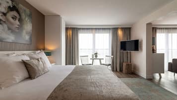 Quarto de hotel com uma cama grande, televisão, uma mesinha com cadeiras e vista para o exterior por grandes janelas.