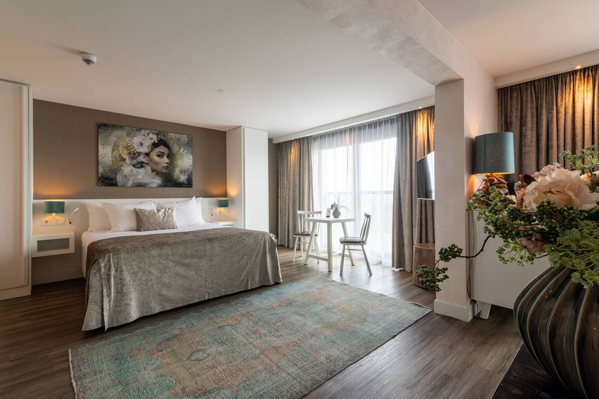Une chambre d’hôtel moderne avec un grand lit, un bureau et une chaise. Un tableau orne le mur et un vase rempli de fleurs est posé sur le bureau.