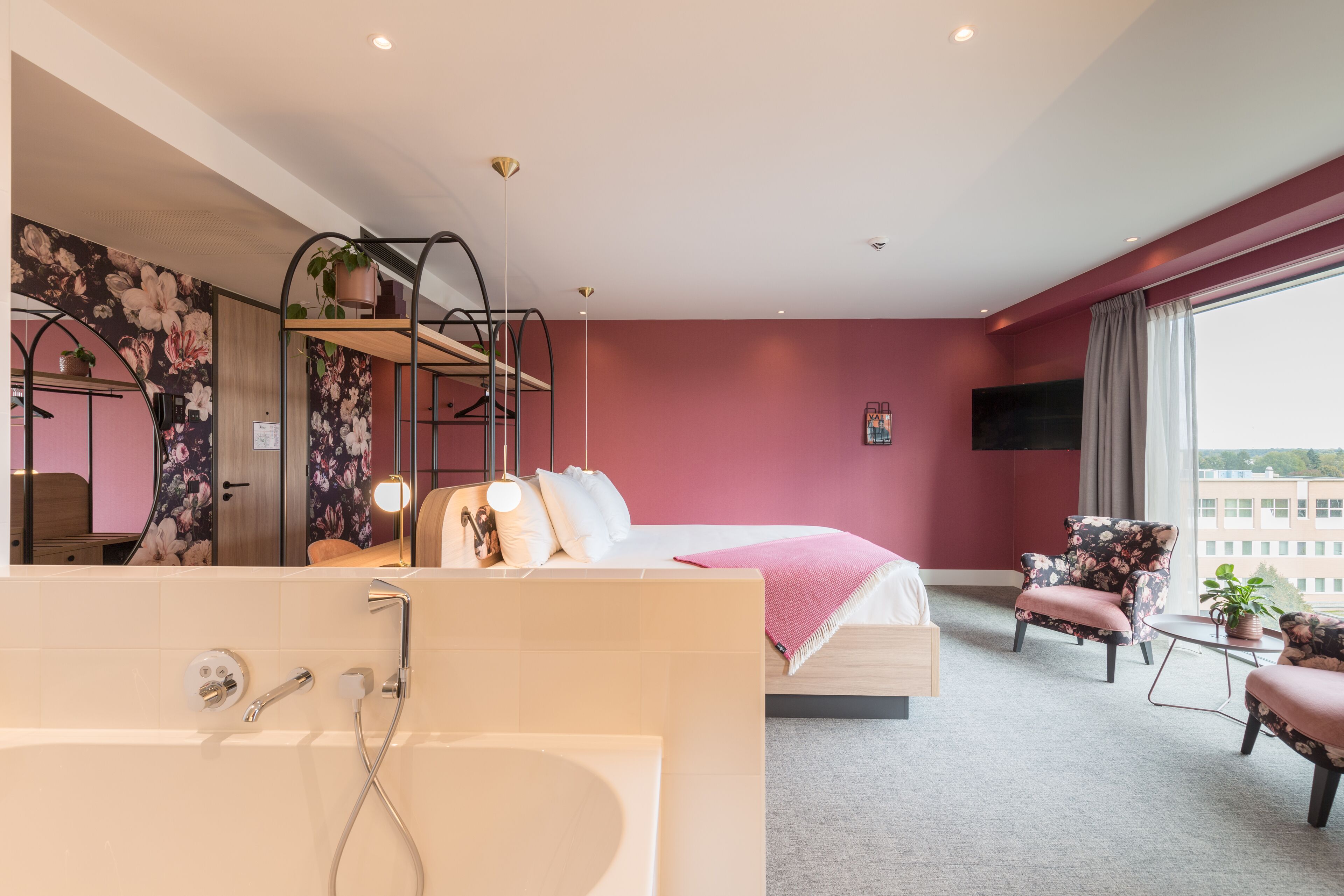 Una habitación de hotel moderna con una cama grande, una zona de estar con una silla, una mesita pequeña y un ventanal con vistas a la ciudad.