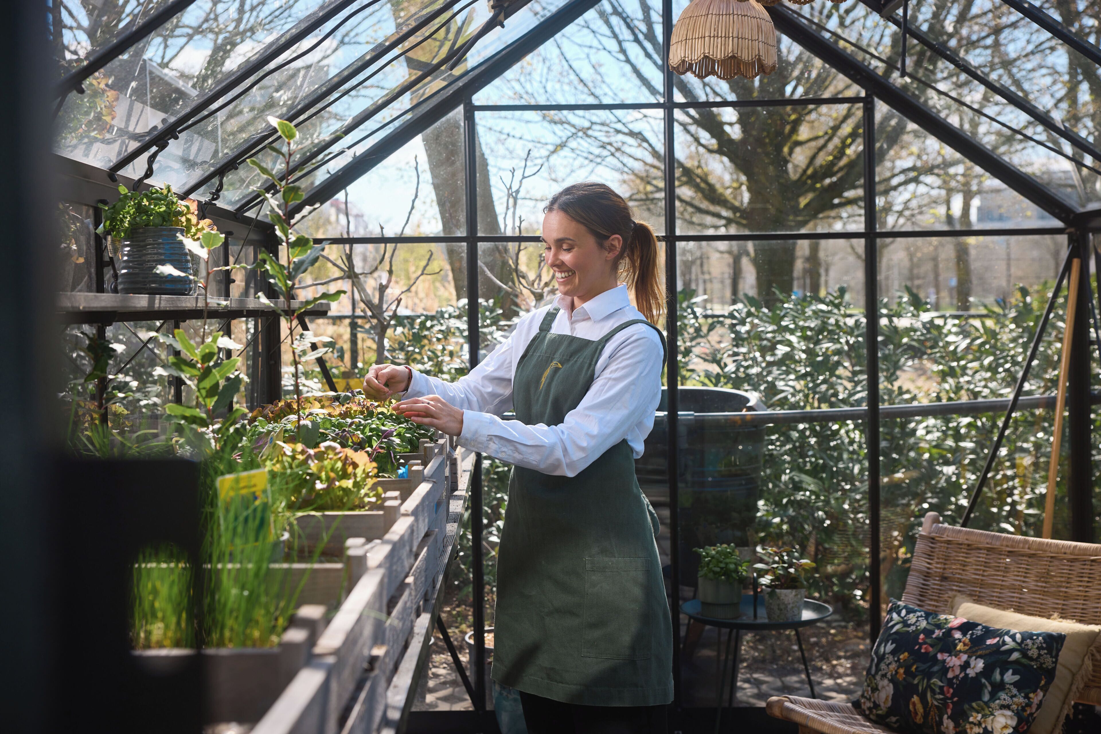 A greenhouse with a person tending to plants, a wicker chair, and a potted plant on a table.