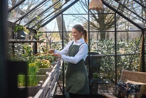 A greenhouse with a person tending to plants, a wicker chair, and a potted plant on a table.