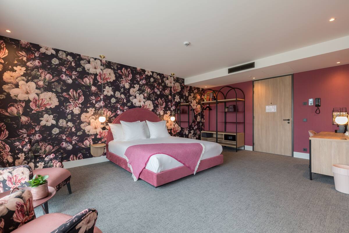 Une chambre d’hôtel avec un papier peint à motifs floraux, un lit recouvert d’une courtepointe rose, un bureau avec une lampe et une armoire.