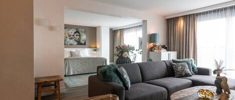 A modern living room with a sofa, a round coffee table, and a bench. There is a painting on the wall and a window with curtains.