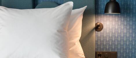 A bedroom with a bed, pillows, a bedside table, a lamp, and a wall with a patterned wallpaper.