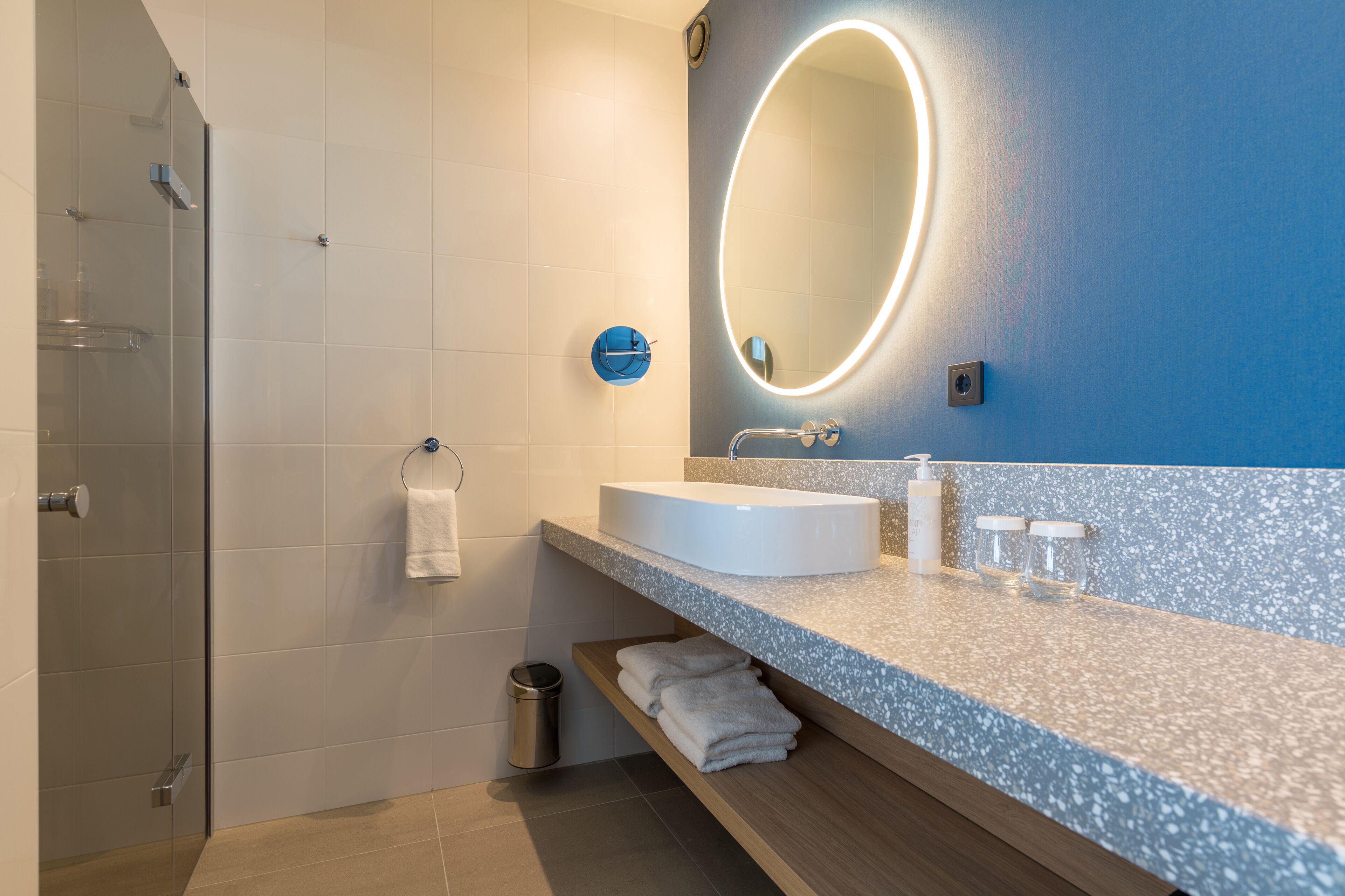 Un baño moderno con cabina de ducha de vidrio, un lavabo blanco, un espejo con luz y un perchero para toallas.