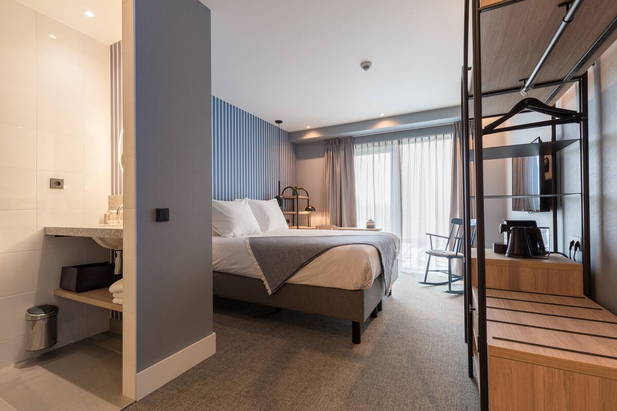 Une chambre d’hôtel moderne équipée d’un lit, d’une table de chevet, d’une chaise, d’un bureau et d’un porte-manteau mural.