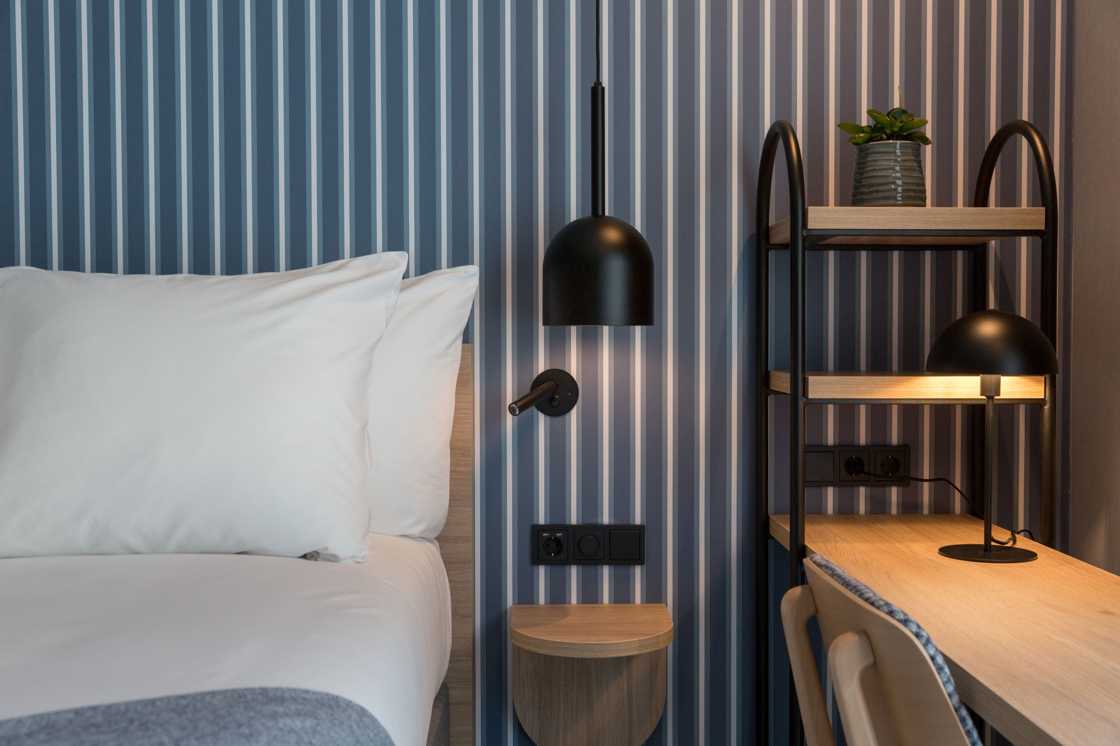 Un dormitorio con una cama, una mesita de noche y una estantería de pared con una lámpara y una planta.