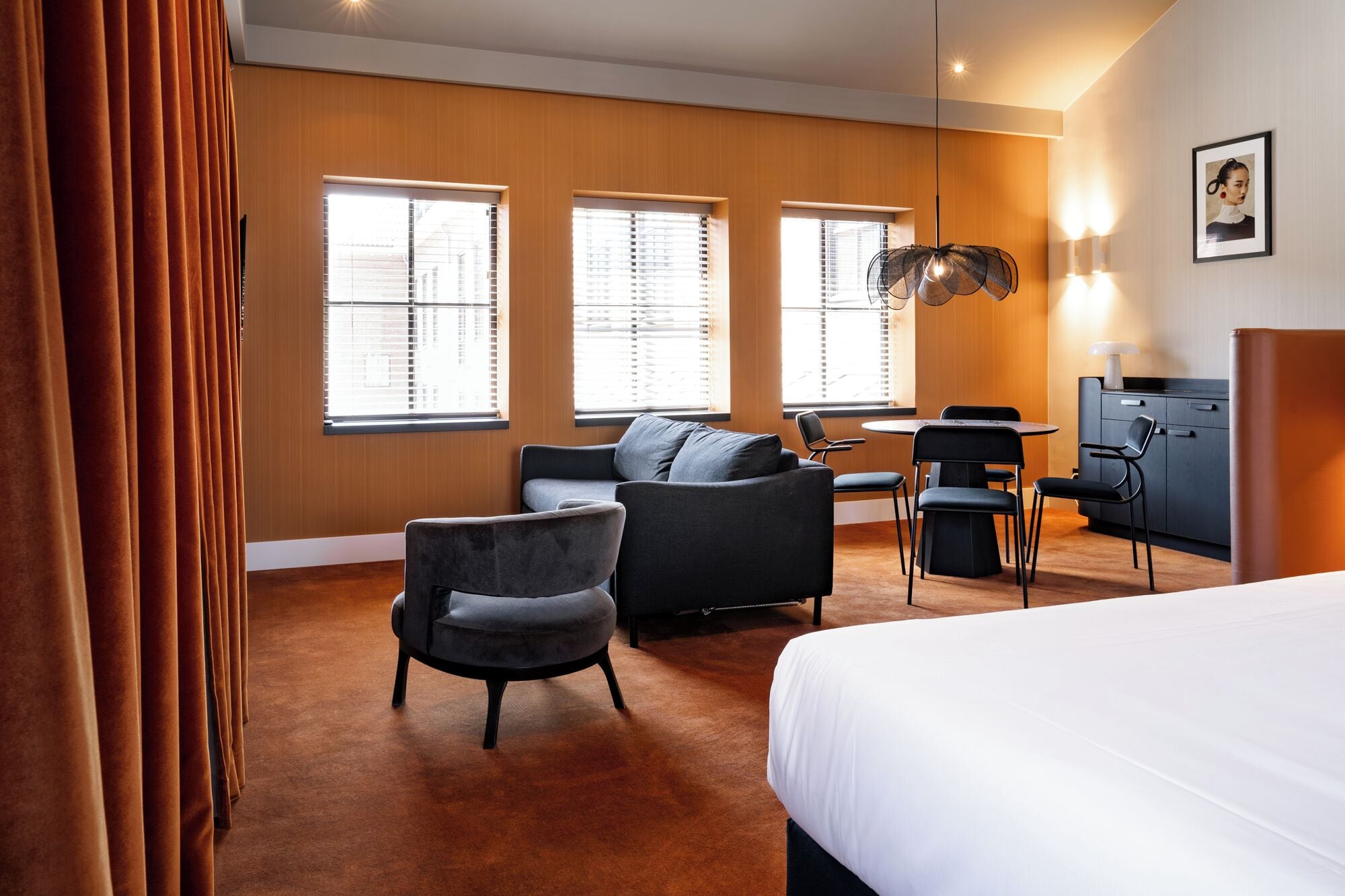 A hotel room with a bed, two armchairs, a desk with a chair, and a wardrobe. There are three windows with blinds, a hanging light fixture, and a framed picture on the wall.
