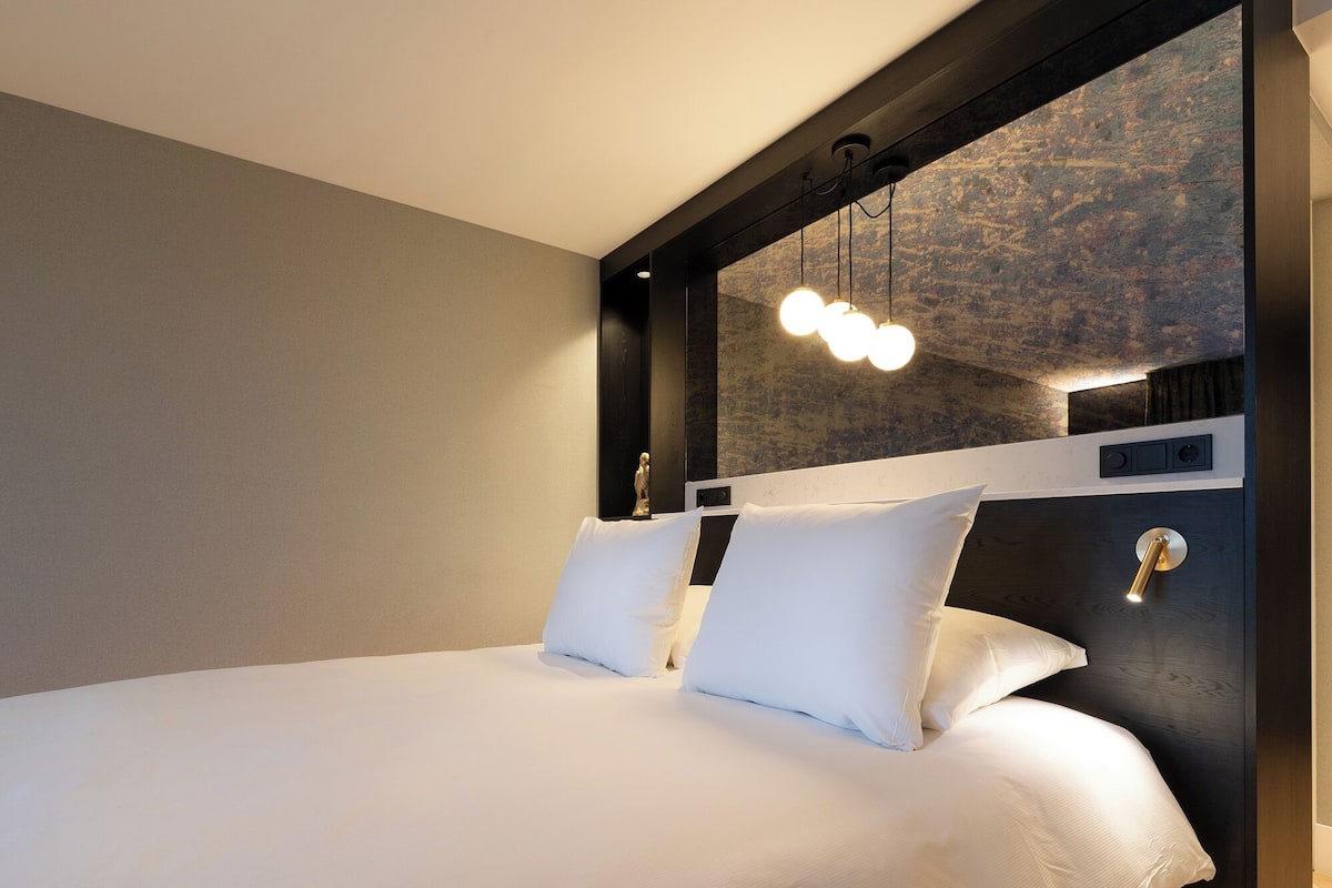 Ein modernes Hotelzimmer mit einem großen Bett, einem Kopfteil mit integrierter Beleuchtung und einer strukturierten Wand dahinter.