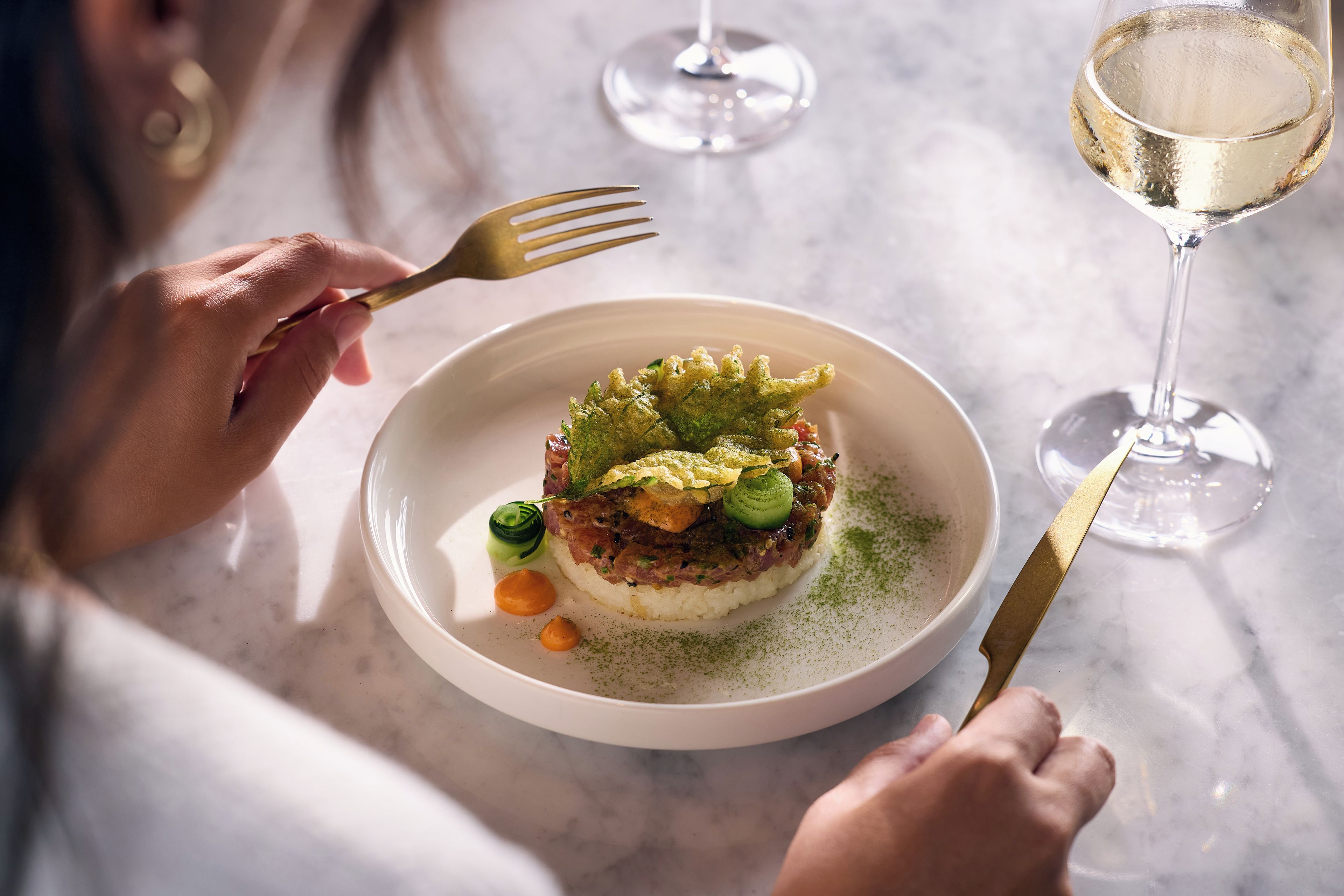 A person dining at a table with a plate of food, a fork, and a glass of white wine.