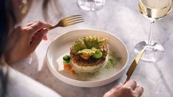 A person dining at a table with a plate of food, a fork, and a glass of white wine.