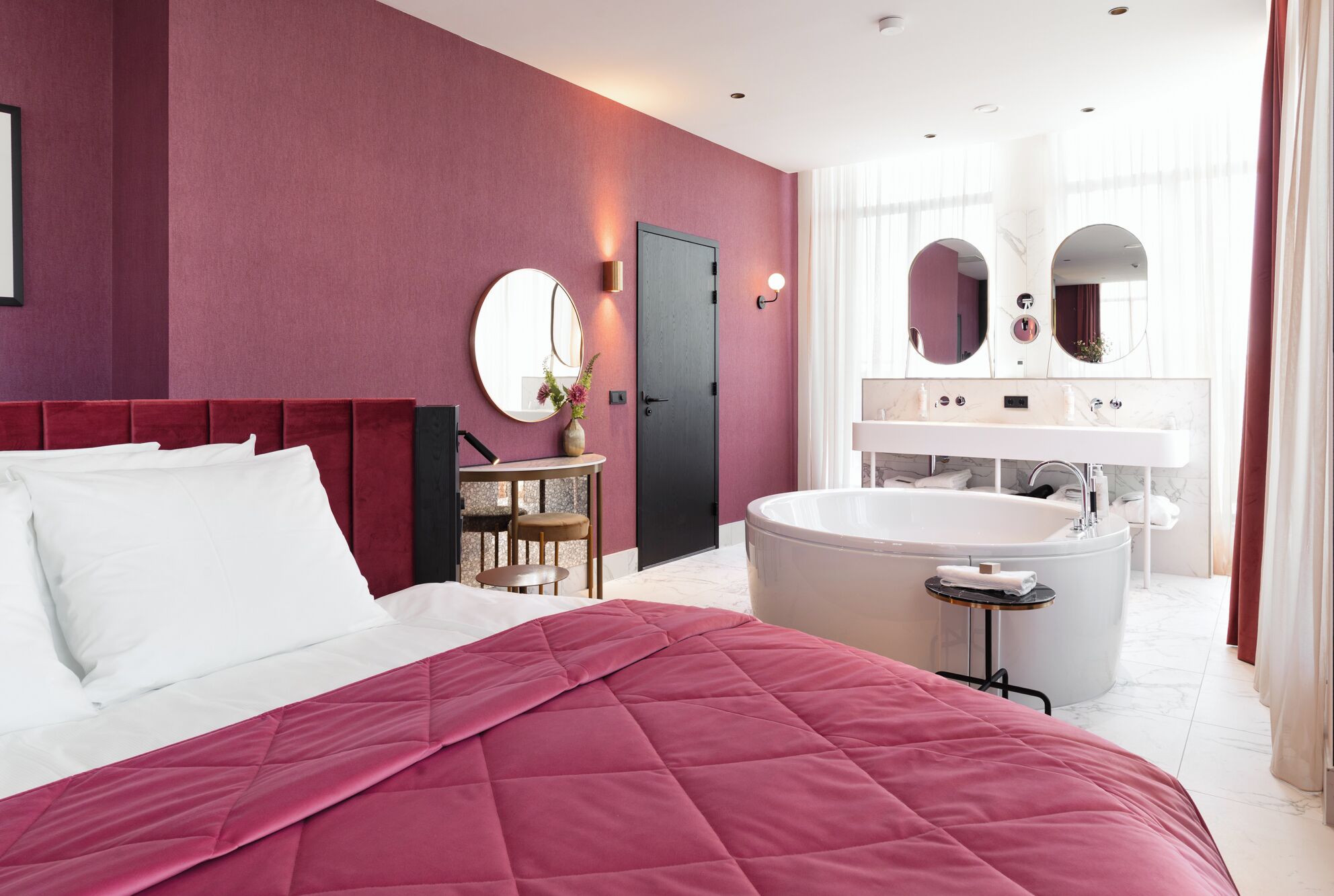 Une chambre d’hôtel moderne dotée d’un grand lit, d’un coin salon avec une table et des chaises, d’une grande baignoire et d’une salle de bain avec un meuble-lavabo et des miroirs.