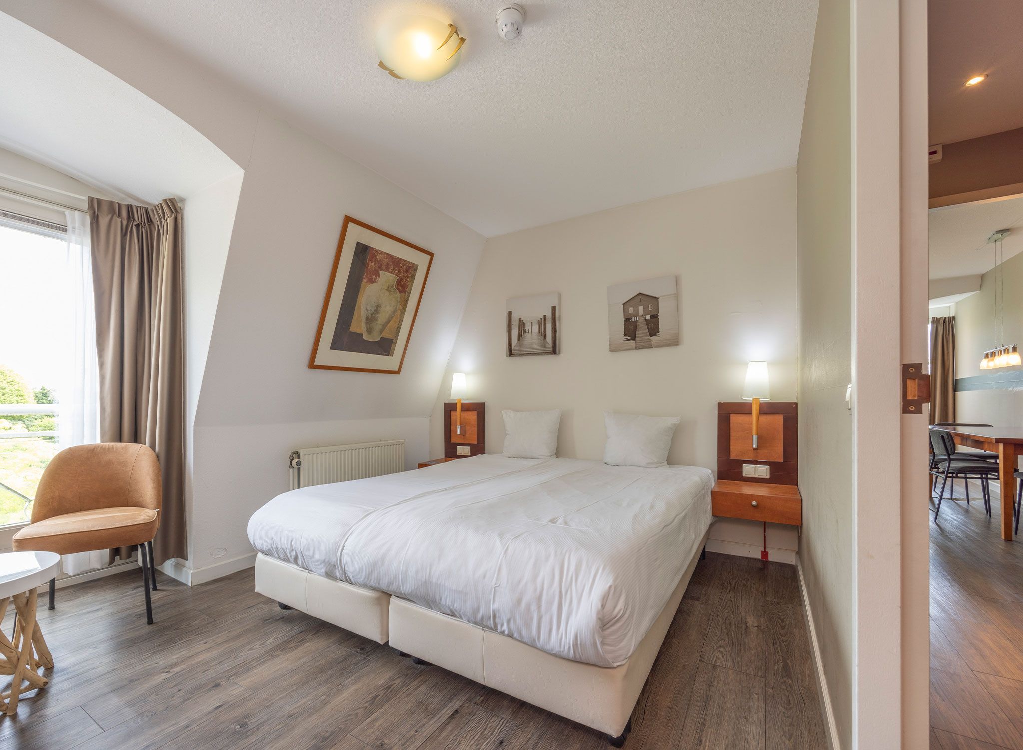 A hotel room with a large bed, a chair, a table, and artwork on the walls.