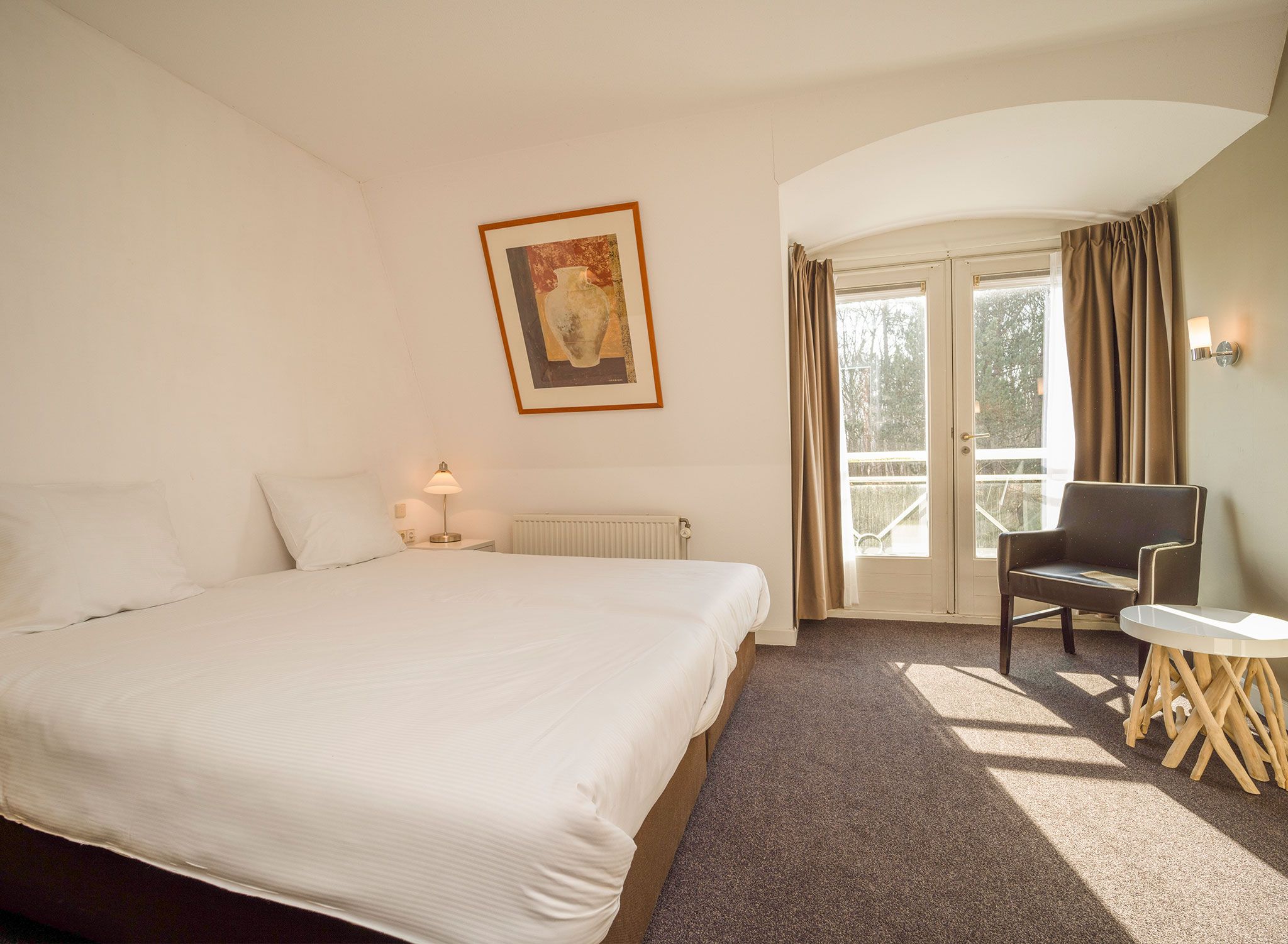 Habitación de hotel con una cama grande, una silla, una mesita, una lámpara y un cuadro en la pared.
