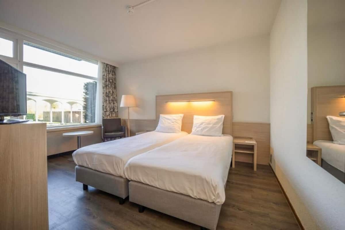 Hotellihuone, jossa on parivuode, työpöytä, tuoli, lamppu ja ikkuna, josta on näkymä.