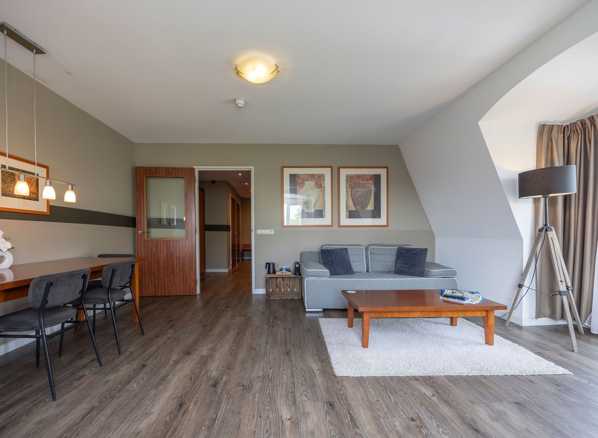 A modern living room with a sofa, coffee table, and chairs. There is a dining area with a table and chairs. The room has wooden flooring and a ceiling light.