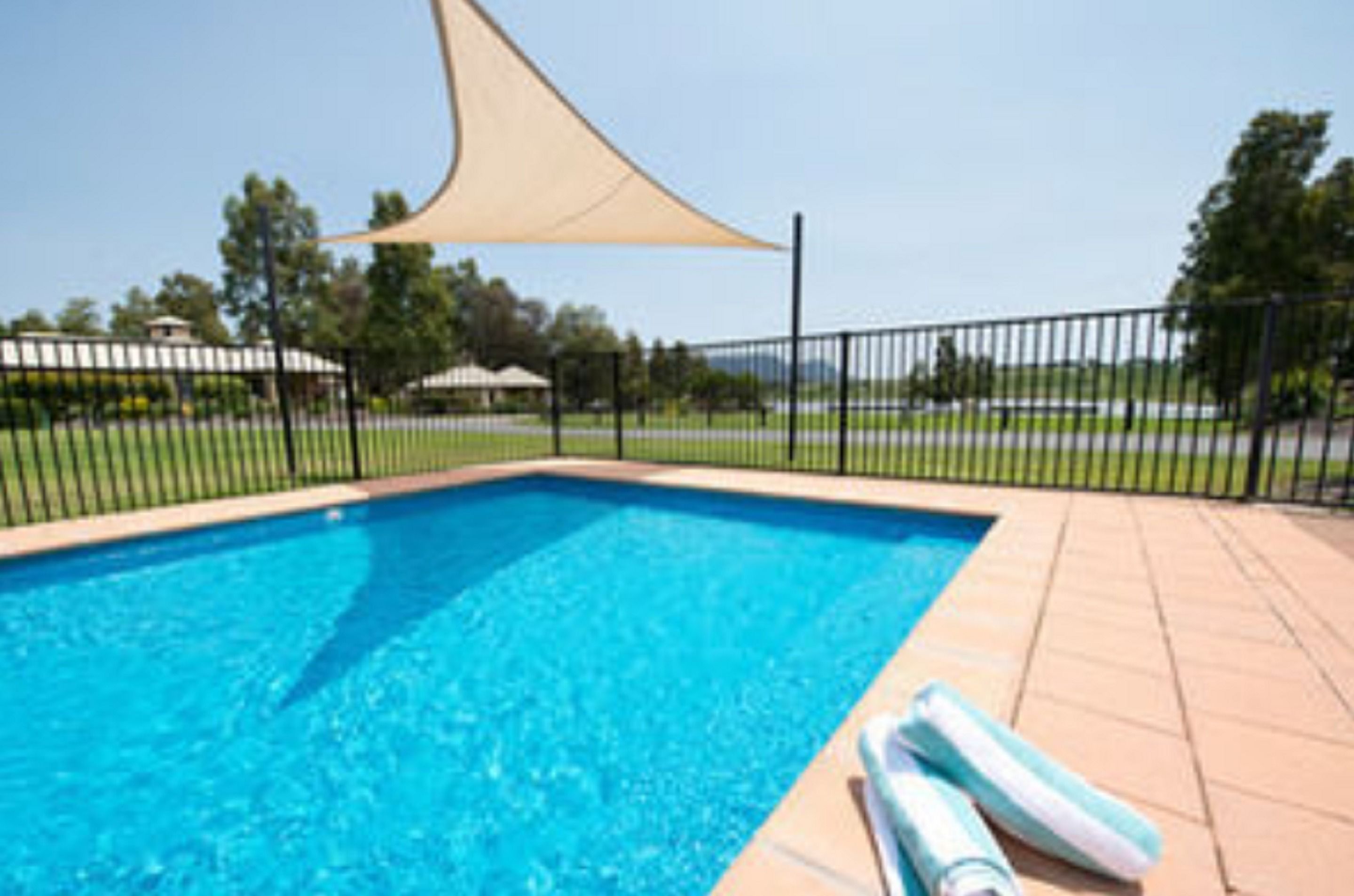 A swimming pool with a tan shade sail, a black metal fence, and a tiled deck.